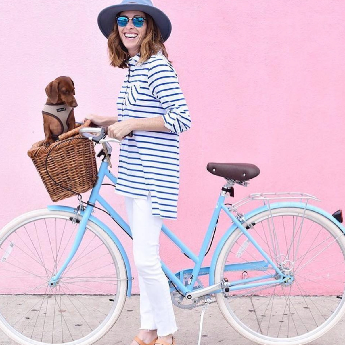A woman holding a blue bicycle with a wicker bike basket with a dachshund inside in front of a pink wall